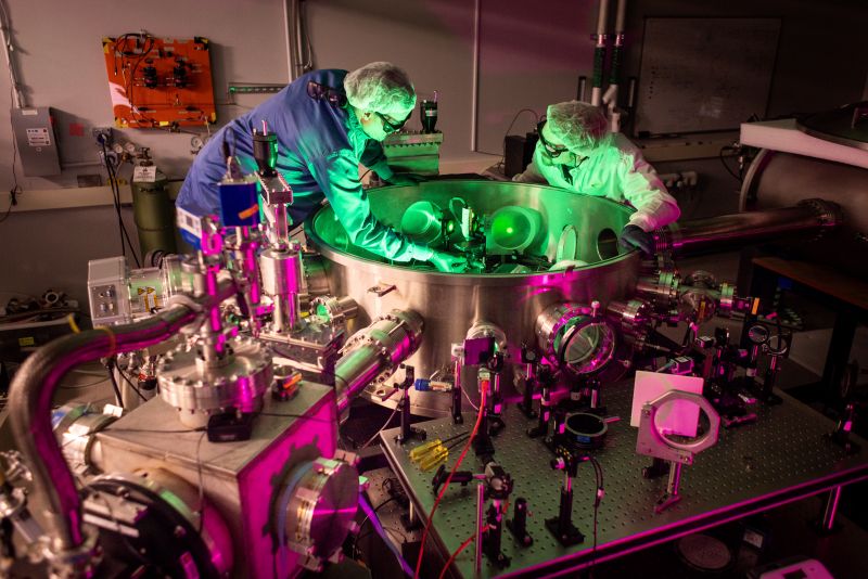 Two men in lab coats, hair nets, gloves and protective eyeglasses adjust the optics inside a circular metal tank. Metal tubes enter the tank from the sides, and more optical components are secured to a table outside.