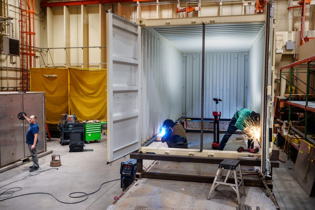 Two technologists weld the Mobile Vault frame to the exterior Conex container while an engineer performs a 3D scan of the vault.